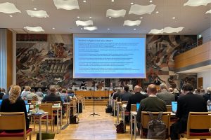 Die URAK-Vorsitzende Heide Glaesmer spricht vor den Synodalen an einem Rednerpult. Zu sehen ist ihre Präsentation zur Arbeit der Aufarbeitungskommission. In der Mitte sitzen mehrere Personen an einem Tisch (Podium). An der Wand hinter ihr und der Leinwand ist ein Flächendeckendes ein modernes Kunstwerk zu sehen.