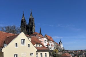 Blick auf die Stadt Meißen mit ihrem Dom mit einem hellen Haus im Vordergrund und weiteren Häusern mit roten Dächern im Hintergrund.
